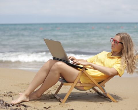 mujer en una silla de playa en la arena frente al mar revisa su computador portatil