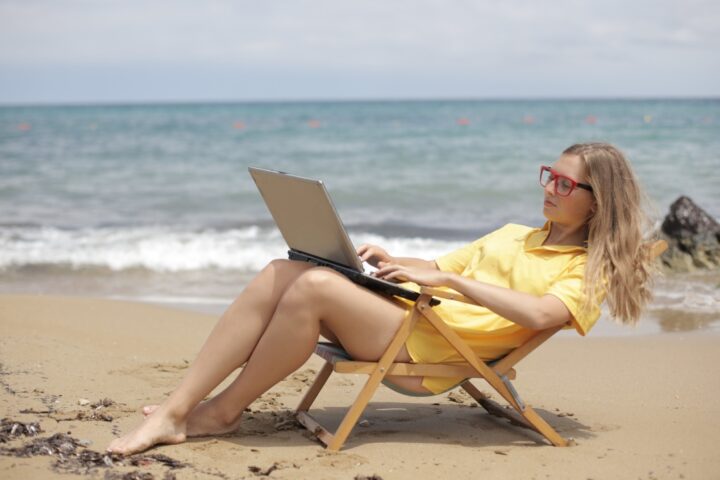 mujer en una silla de playa en la arena frente al mar revisa su computador portatil